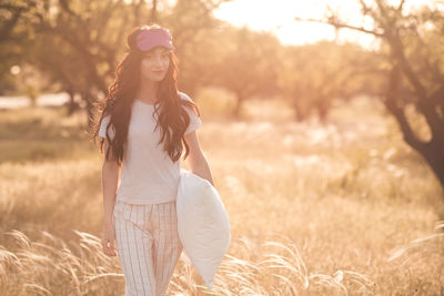 Pretty young woman with face mask wearing pajamas and holding pillow in meadow outdoors. 