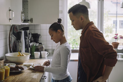 Side view of father looking at daughter preparing breakfast while standing in kitchen at home