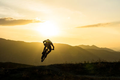 Rear view of man with bicycle performing stunt on mountain against sky during sunset