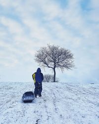 Man with sled walking on snow covered field against sky during winter