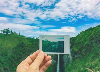 Cropped hand of person holding instant print transfer against waterfall and mountain