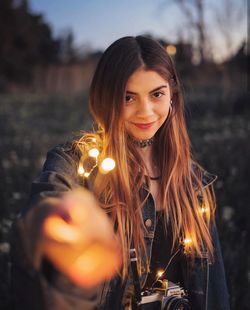 Portrait of smiling young woman holding illuminated string lights while standing on land during sunset