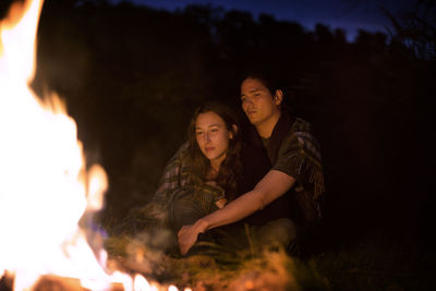Couple wrapped in blanket sitting by campfire at night