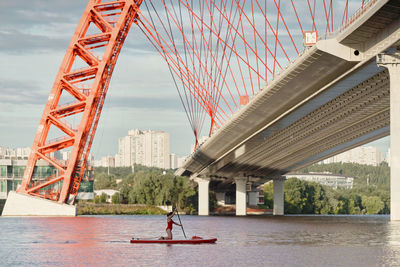 Side view of woman standing on paddle board in river and rowing with paddle under bridge in city