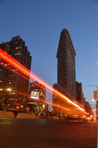 Light trails on city street by buildings against sky at night