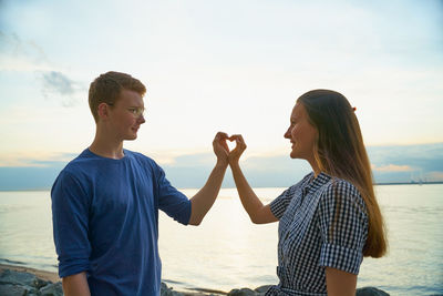 Young couple making heart shape by hand while standing at beach against sky