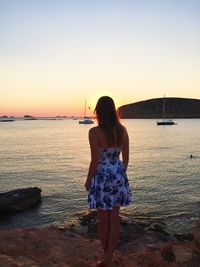 Rear view of young woman standing on rocks by sea against sky during sunset