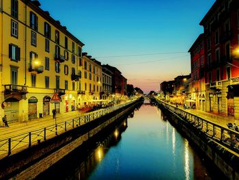 Canal amidst illuminated city against sky at sunset