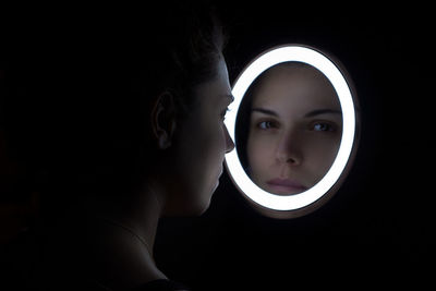 Close-up of young woman looking through mirror reflection over black background