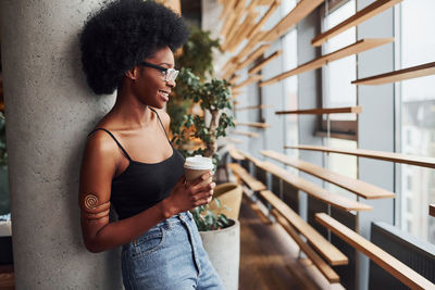 African girl with curly black hair and in casual clothes standing indoors with cup of drink.