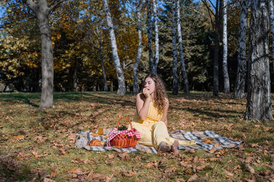 Beautiful caucasian woman in yellow dress on a picnic outdoors, sitting on a plaid in forest