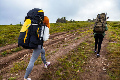 Two hiking women with backpack go along the path to the mountains 