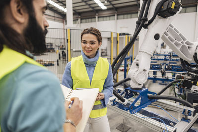 Smiling engineer looking at colleague with note pad standing by robotic arm