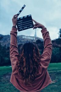 Rear view of woman with arms raised holding film slate against sky