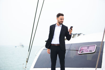 Businessman using phone while standing on boat in sea against clear sky