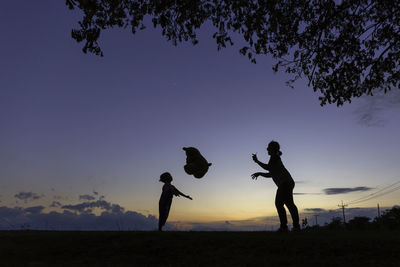 Silhouette woman throwing teddy bear to daughter on field against sky during sunset