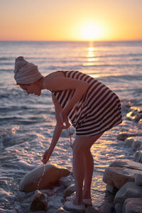 Full length of woman on rock at beach against sky during sunset