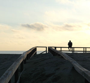 Man standing on railing by sea against sky during sunset