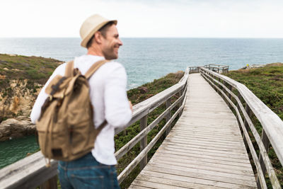 Back view of active travelling man in casual outfit and hat with backpack exploring nature while walking along wooden walkway in countryside