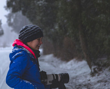 Side view of woman wearing warm clothing while holding digital camera in forest during snowfall