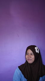 Smiling young woman wearing hijab looking away against purple wall