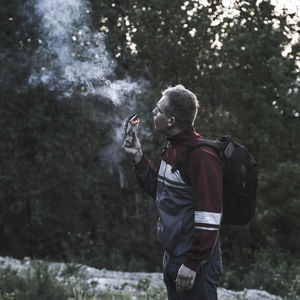 Side view of man with backpack holding burning paper while standing in forest