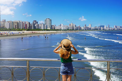 Traveler girl holding hat enjoying view of skyscrapers skyline and waving ocean of santos, brazil