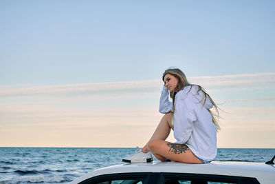 Side view of serene female traveler sitting on car roof and enjoying sunset in evening on background of sea in summer during vacation