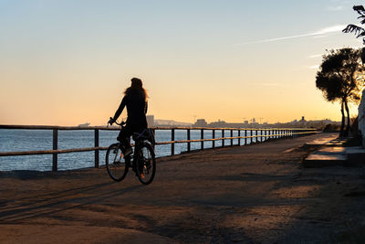 Silhouette woman riding bicycle on promenade against sky during sunset