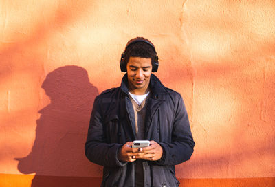 Young man using mobile phone while standing against wall
