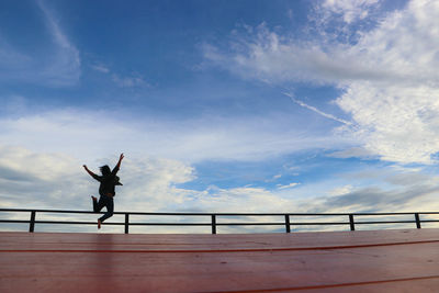 Silhouette young man with arms raised jumping on footbridge against sky