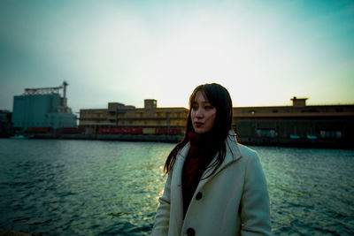 Young woman standing by river against sky during sunset