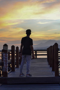 Rear view of man standing on railing against sky during sunset