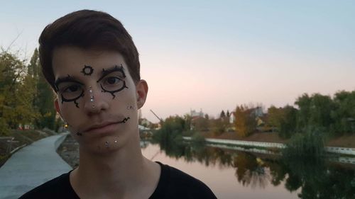 Close-up portrait of young man with face paint standing by lake against sky