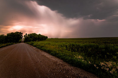 Empty road by field against storm clouds during sunset