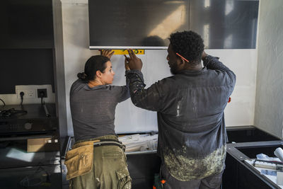 Rear view of male and female carpenters measuring cabinet with equipment in kitchen