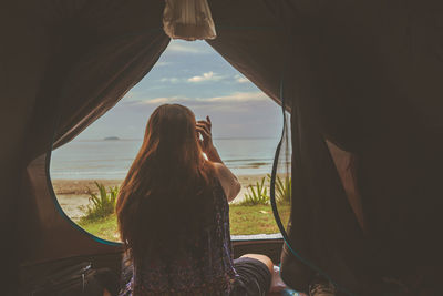 Rear view of woman looking through window at sunset