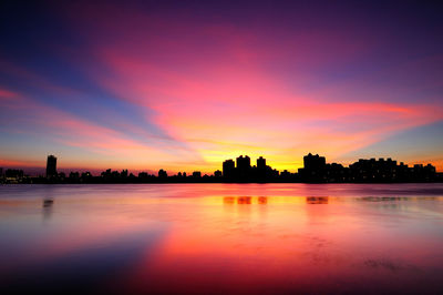 Silhouette buildings by lake against romantic sky at sunset