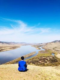 Rear view of man sitting on rock against blue sky