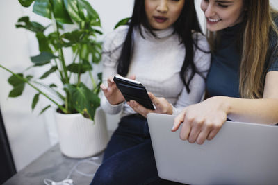 Midsection of female bloggers using smart phone while sitting on sideboard in creative office