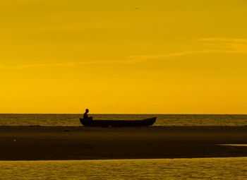 Silhouette man sitting on rowboat at beach against orange sky