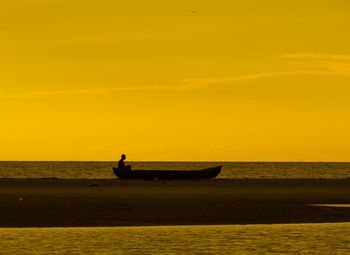 Silhouette man sitting on rowboat at beach against orange sky