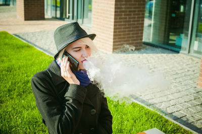 Young woman talking on mobile phone while smoking by built structure