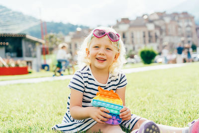 Little happy girl in sunglasses sitting on the lawn with anti-stress pop it smiles. 