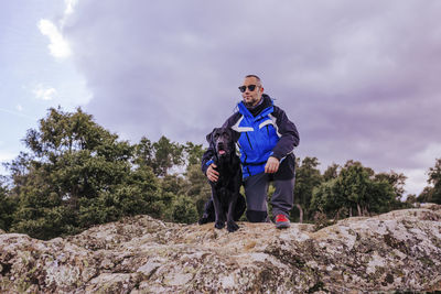 Low angle view of man with dog kneeling on rock against cloudy sky