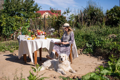 A woman in a hat is sitting on a chair near the table, a labrador retriever is lying next to her