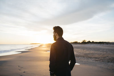 Rear view of mid adult man standing at beach against cloudy sky during sunset