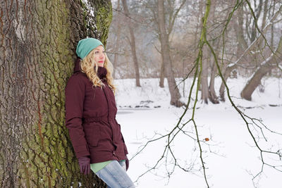 Thoughtful woman leaning on tree trunk in snow covered forest