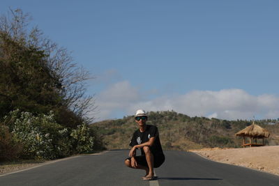 Portrait of young man crouching on road against sky