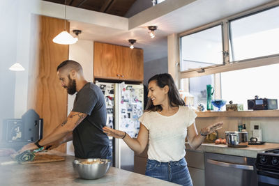 Happy woman gesturing while talking to man cutting vegetables in kitchen